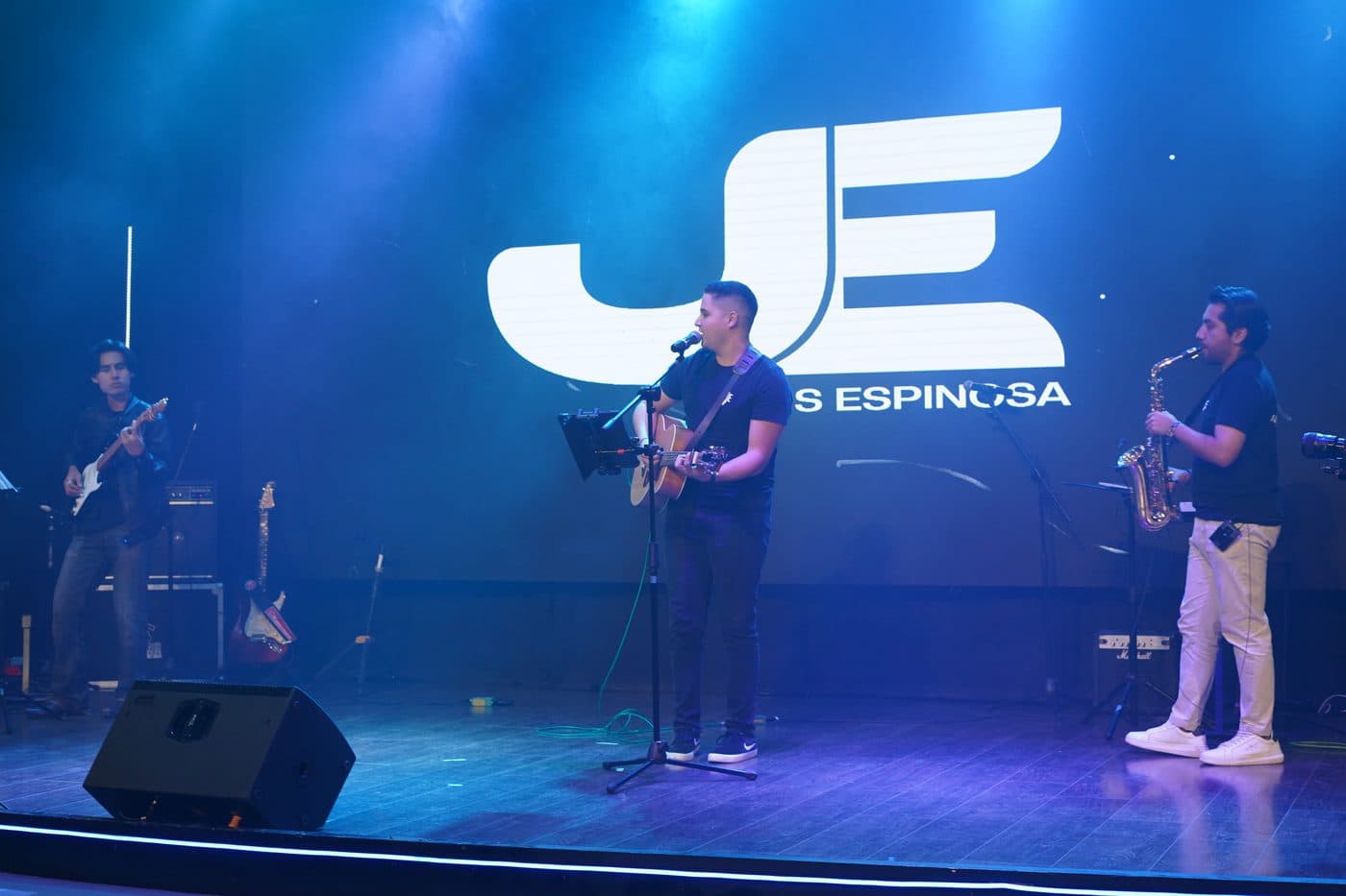 Jesús Espinoza cantando con guitarra junto a un saxofonista, bajo el logotipo JE